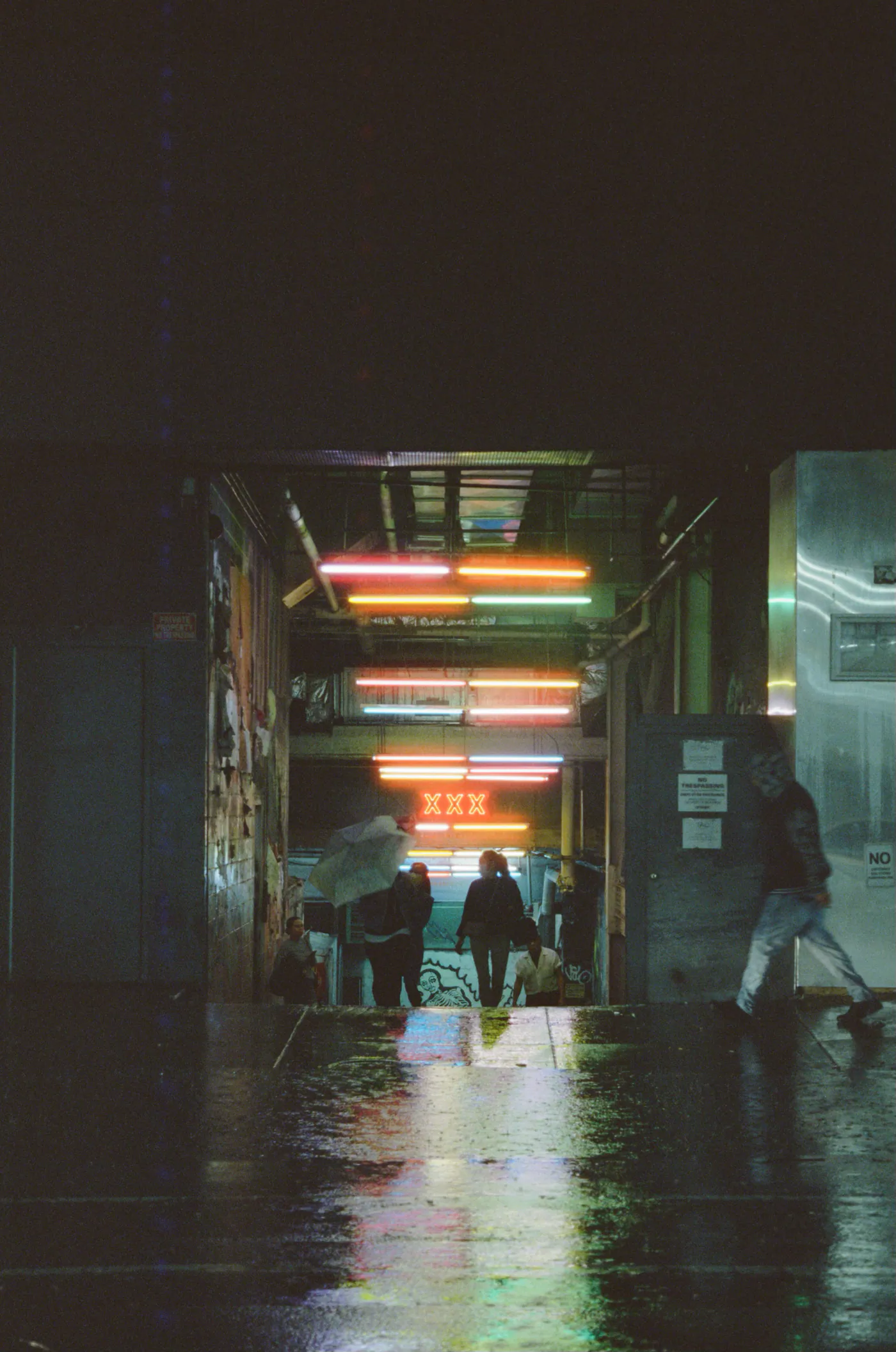 Eingang einer Passage bei Nacht und Regen mit Neonröhren, Menschen darunter, ein Schirm